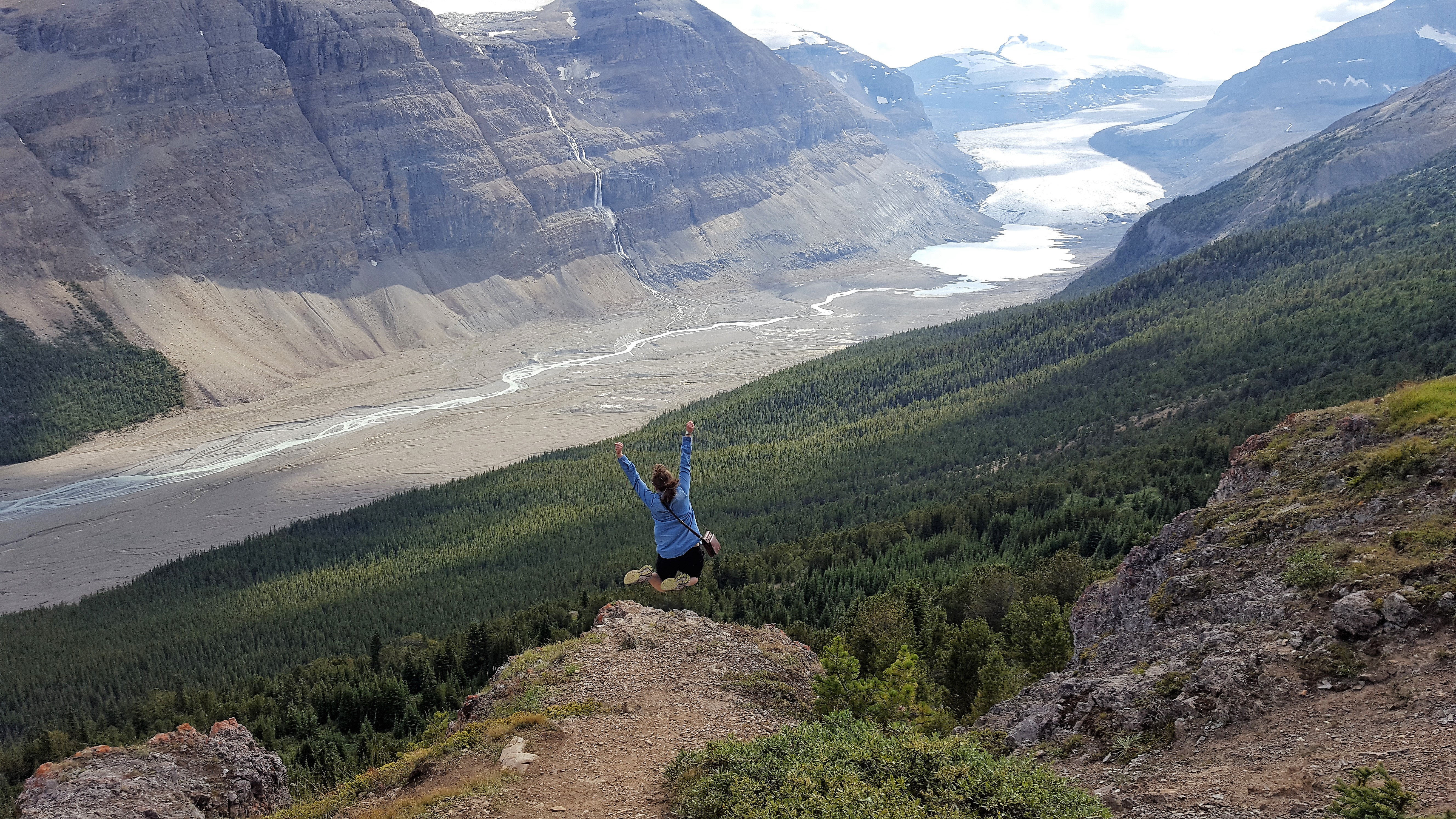 white woman with back to camera mid-jump with hands and arms outstretched above their head and feet tucked up in a jump. They are standing on the edge of a cliff/ridge with a wide vista of a valley in the background, including the Saskatchewan Glacier and mouth of the North Saskatchewan river in the background.